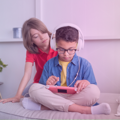 Child looking over the shoulder of another child playing a hand held video gaming device.